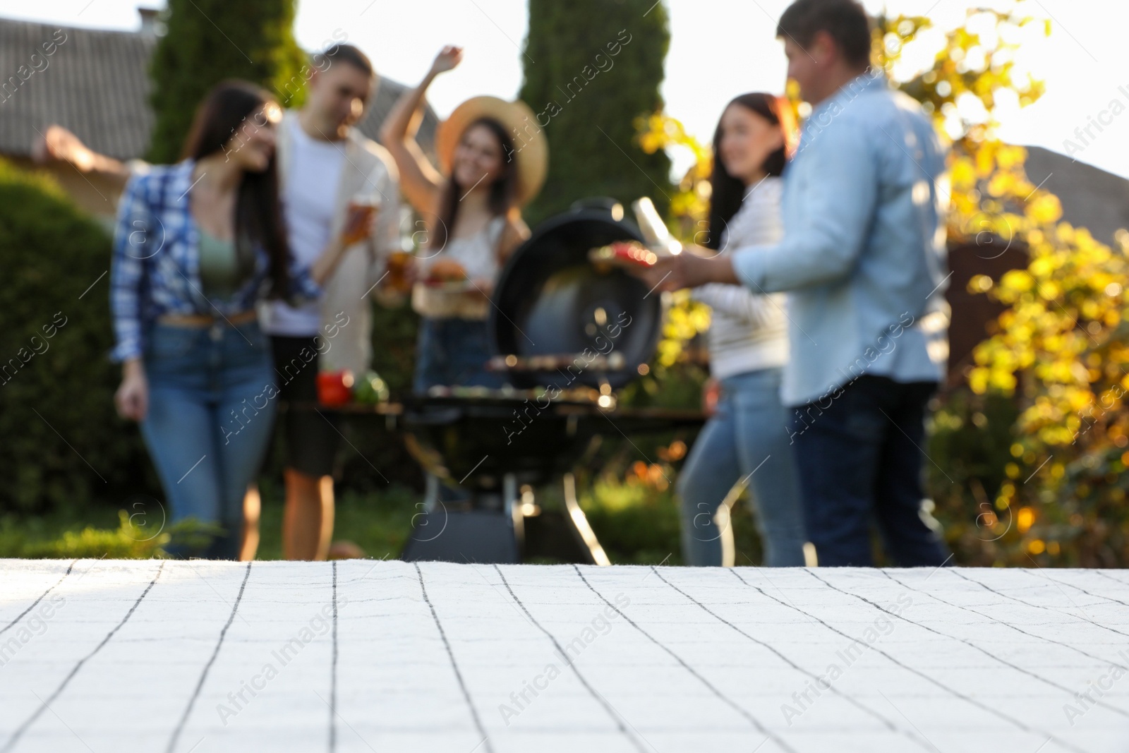 Photo of Empty white wooden table and blurred view of friends having barbecue party outdoors. Space for text