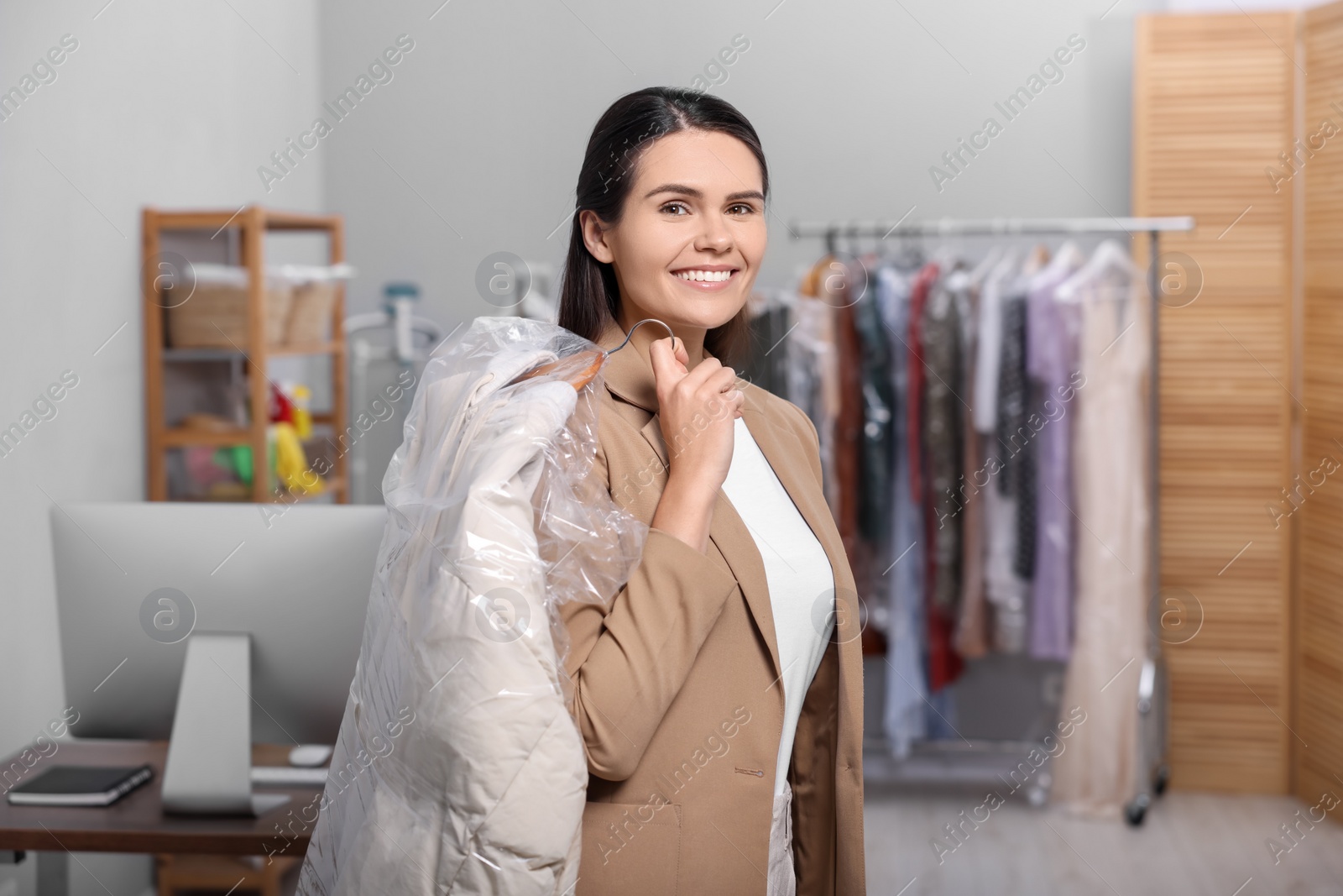 Photo of Dry-cleaning service. Happy woman holding hanger with jacket in plastic bag indoors