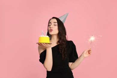Coming of age party - 21st birthday. Woman holding delicious cake and sparkler and blowing number shaped candles on pink background