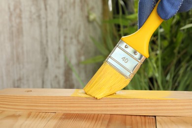 Photo of Worker applying yellow paint onto wooden surface against blurred background, closeup. Space for text