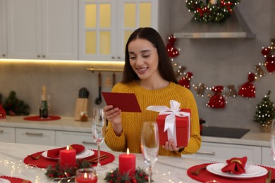 Happy young woman with Christmas gift reading greeting card at table in kitchen