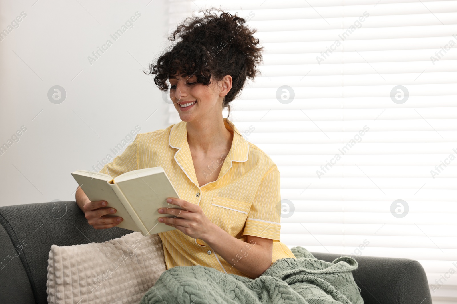 Photo of Beautiful young woman in stylish pyjama reading book on sofa at home