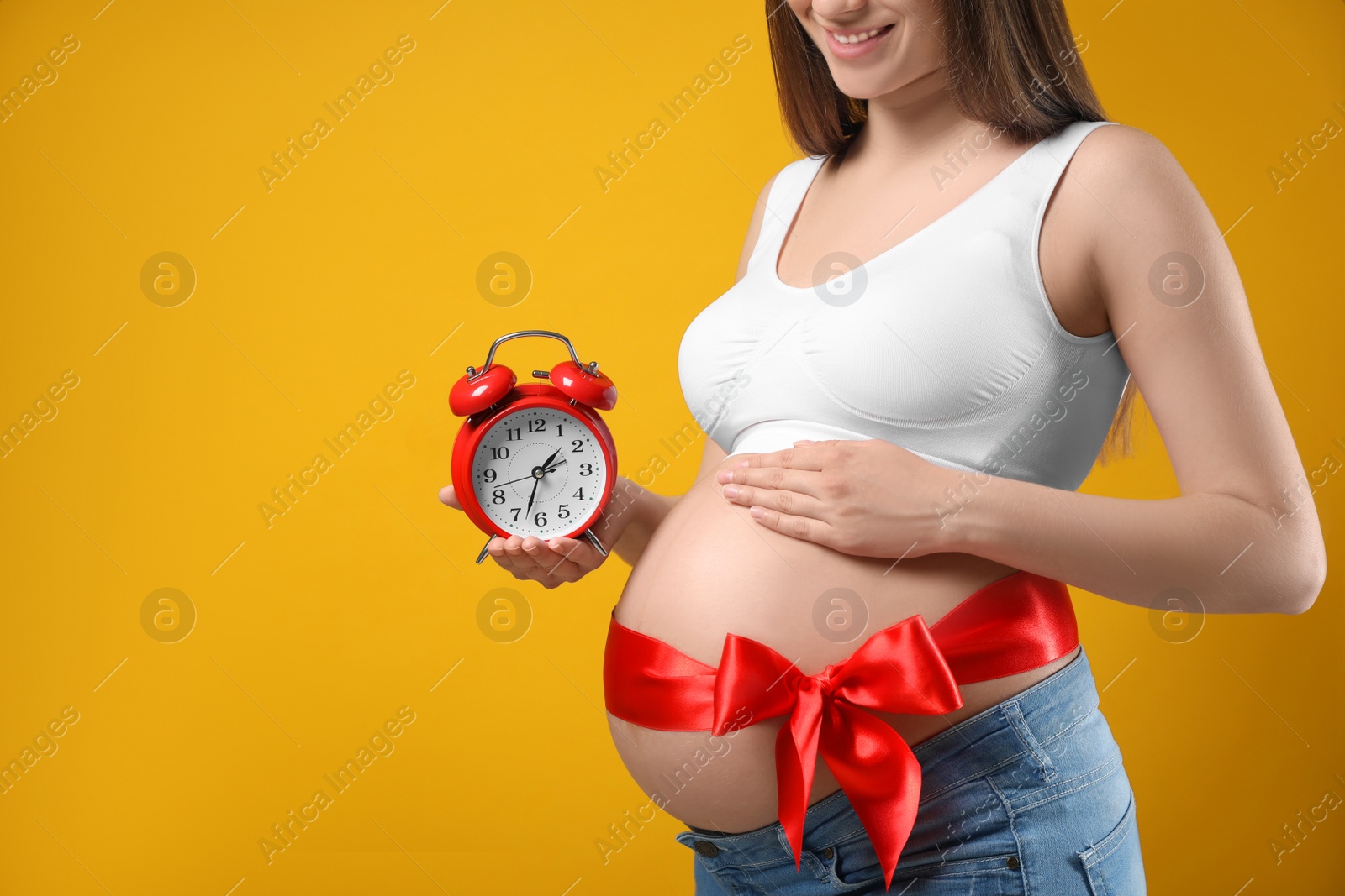 Photo of Young pregnant woman with alarm clock and red bow on yellow background, closeup. Time to give birth