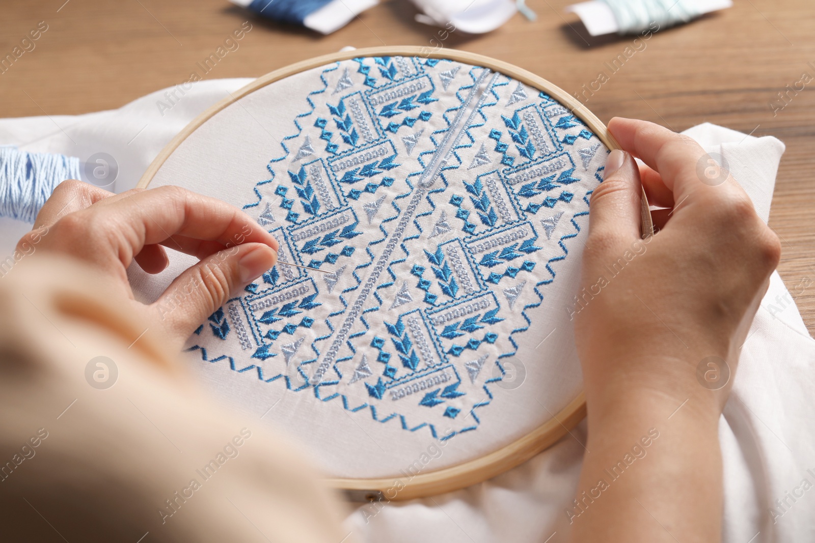 Photo of Woman embroidering white shirt with blue thread at wooden table, closeup. Ukrainian national clothes