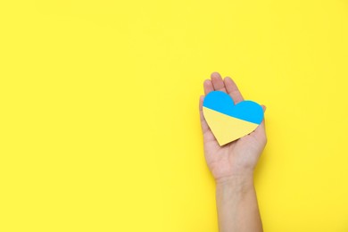 Photo of Woman holding paper heart in colors of Ukrainian flag on yellow background, top view. Space for text