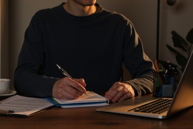 Photo of Man taking notes at wooden table in office in evening, closeup