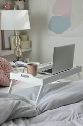 White tray table with laptop and cup of drink on bed indoors
