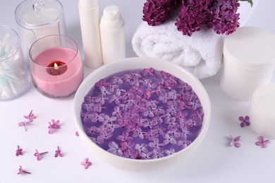 Photo of Bowl of water with lilac flowers, spa products and burning candle on white table