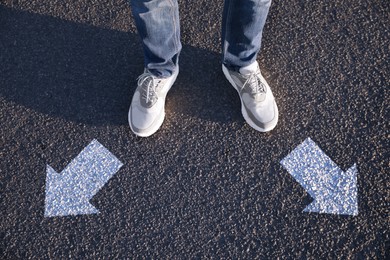 Image of Choice of way. Man standing in front of drawn marks on road, closeup. White arrows pointing in different directions