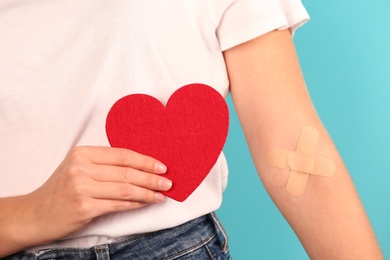 Woman holding heart near hand with adhesive plasters against color background, closeup. Blood donation