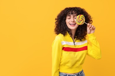 Beautiful woman covering eye with lollipop on yellow background, space for text