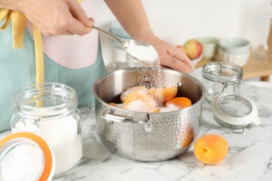 Woman adding sugar into pot with apricots at table in kitchen, closeup. Making delicious jam