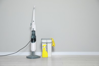 Modern steam mop, bucket with gloves and spray of cleaning product on floor near grey wall, space for text