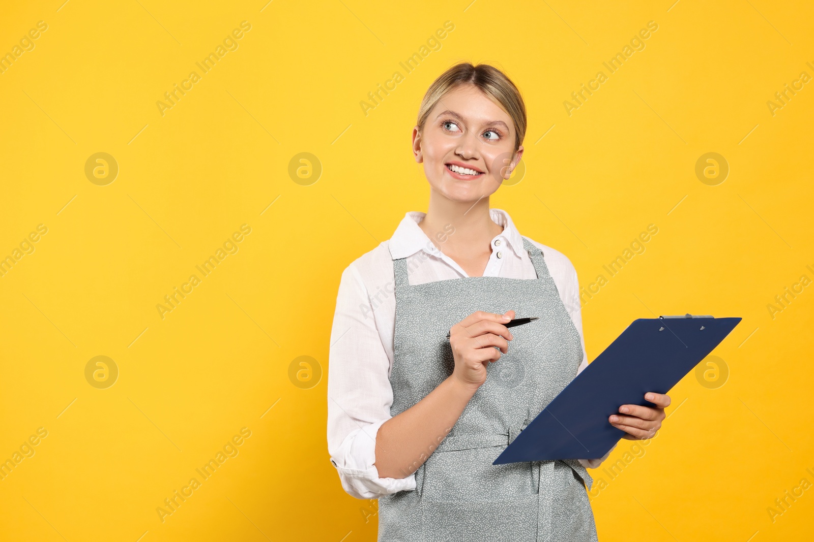 Photo of Beautiful young woman in clean apron with clipboard on orange background. Space for text