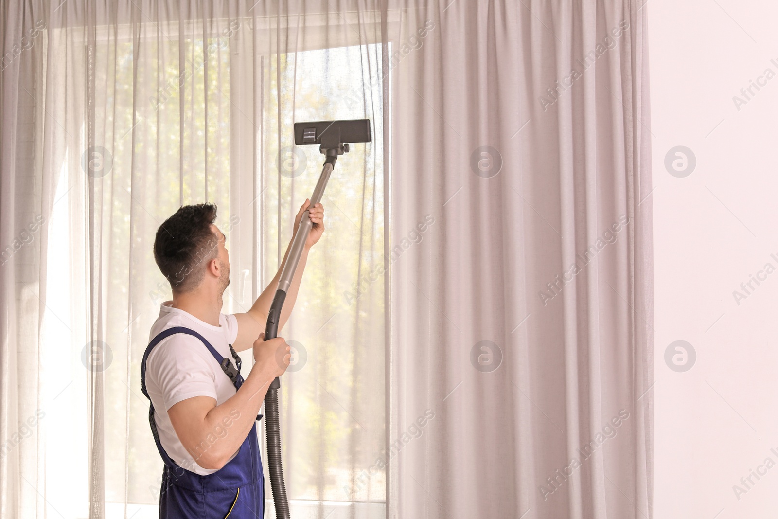 Photo of Male worker removing dust from curtains with professional vacuum cleaner indoors