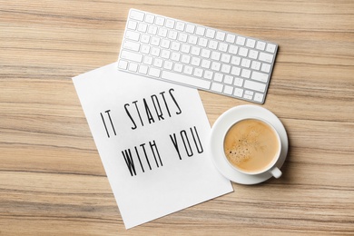Photo of Sheet of paper with phrase It Starts With You, coffee and keyboard on wooden table, flat lay