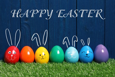 Several eggs with drawn faces and ears as Easter bunnies among others on green grass against blue wooden background