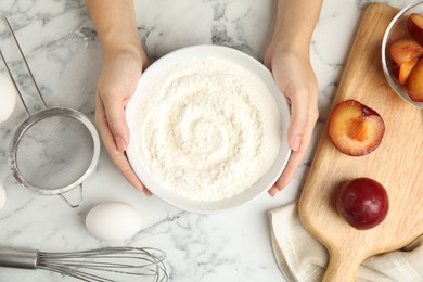 Woman holding bowl with flour at white marble table, top view. Cooking of delicious plum cake