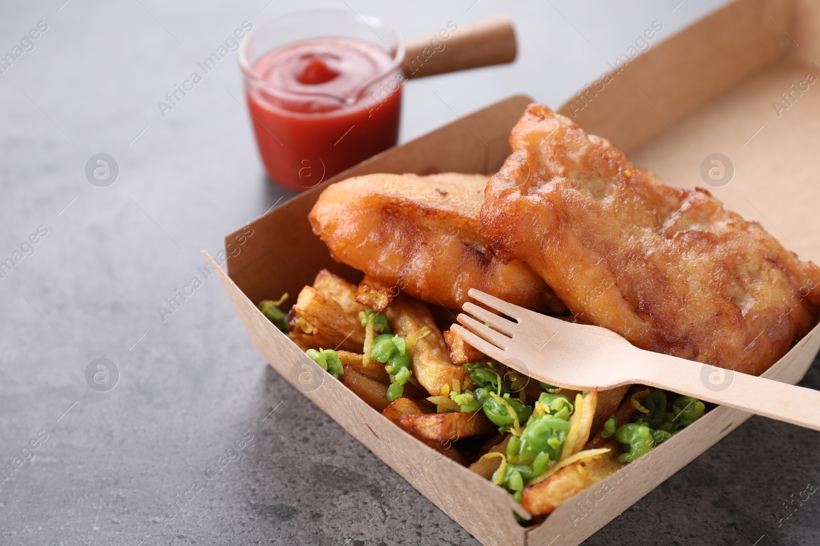 Photo of Tasty fish, chips, sauce and peas in paper box on grey table, closeup. Space for text