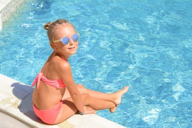 Happy cute little girl sitting at edge of swimming pool on sunny day. Space for text