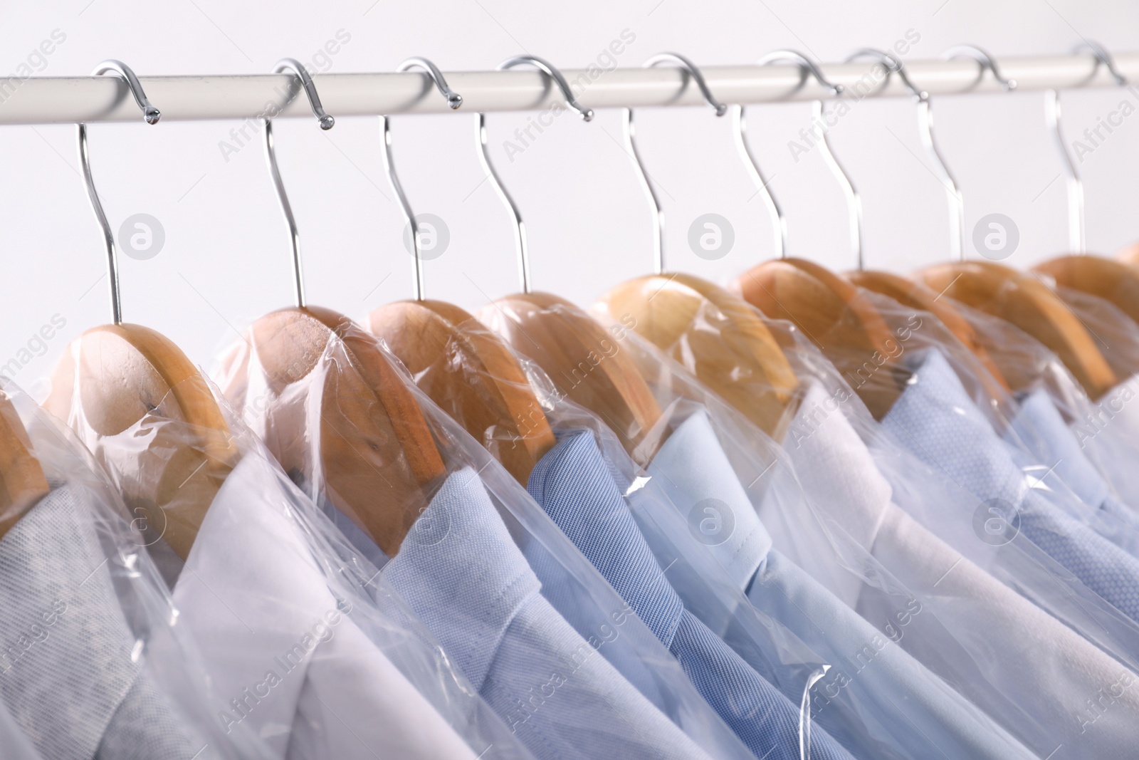 Photo of Hangers with shirts in dry cleaning plastic bags on rack against light background, closeup