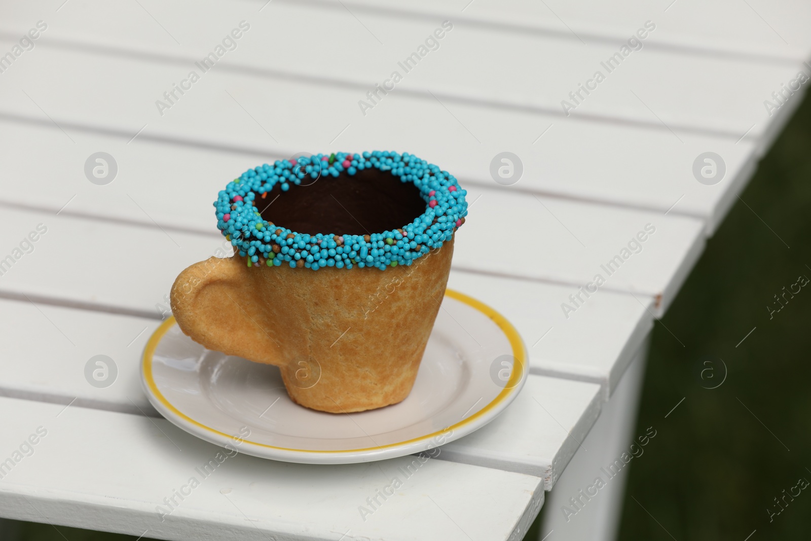 Photo of Delicious edible biscuit coffee cup decorated with sprinkles on white wooden table outdoors