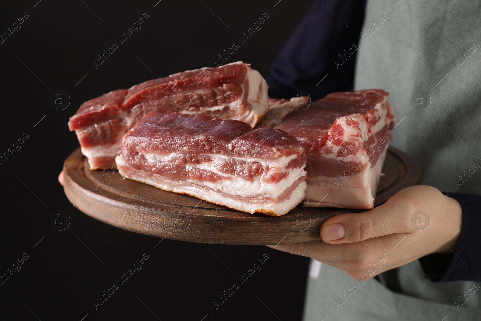 Photo of Woman holding wooden board with pieces of raw pork belly on black background, closeup