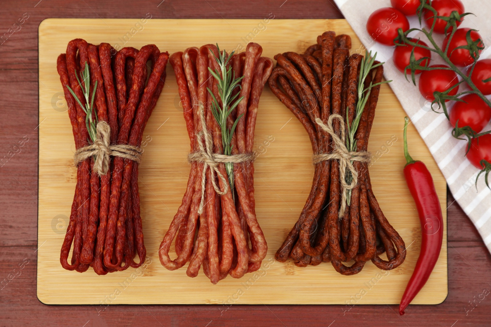 Photo of Bundles of delicious kabanosy with rosemary, tomatoes and chilli on wooden table, flat lay