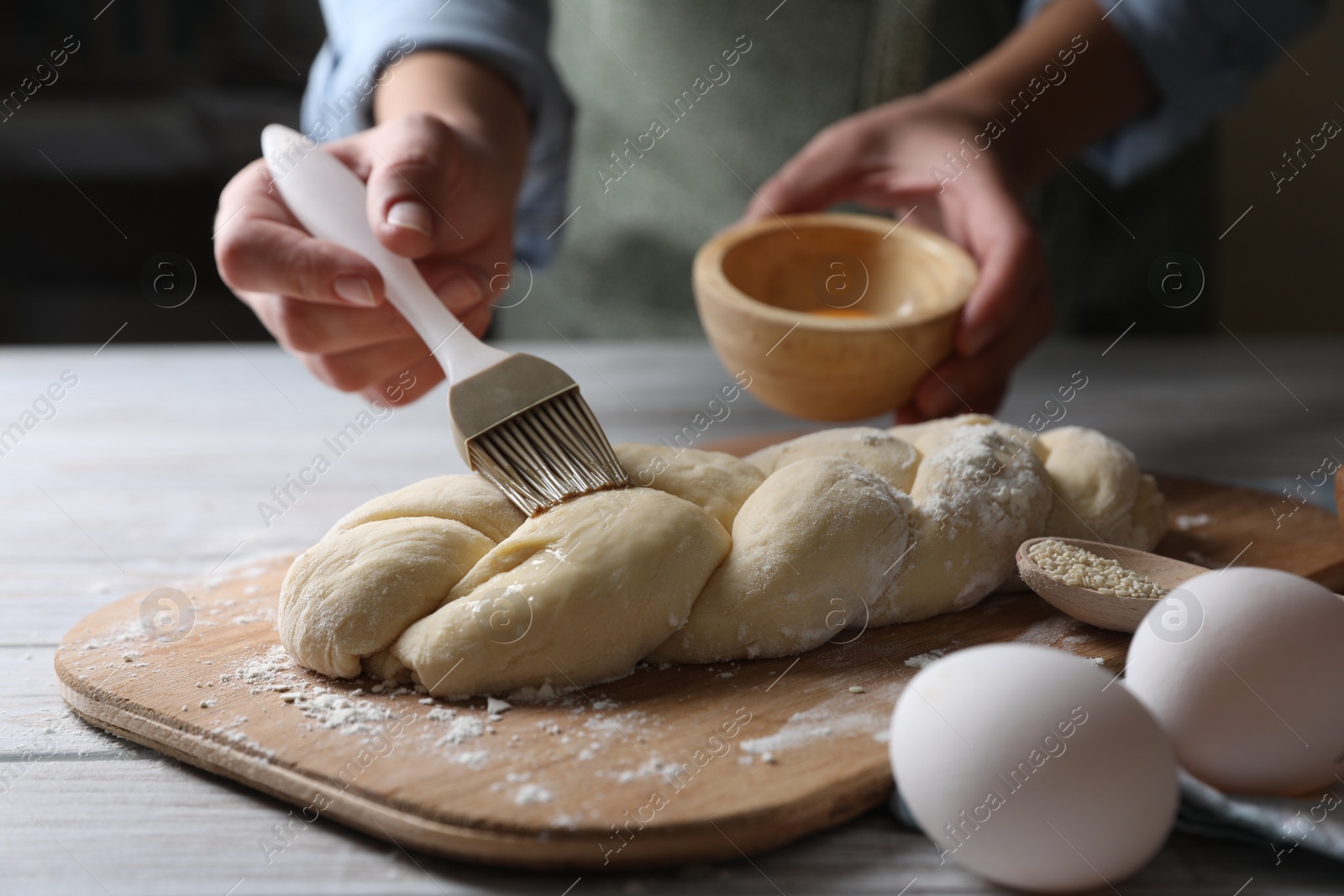 Photo of Woman spreading egg yolk onto raw braided bread at white wooden table, closeup. Traditional Shabbat challah