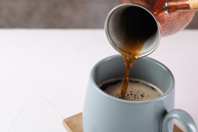 Photo of Pouring aromatic coffee from cezve into cup at white table, closeup. Space for text