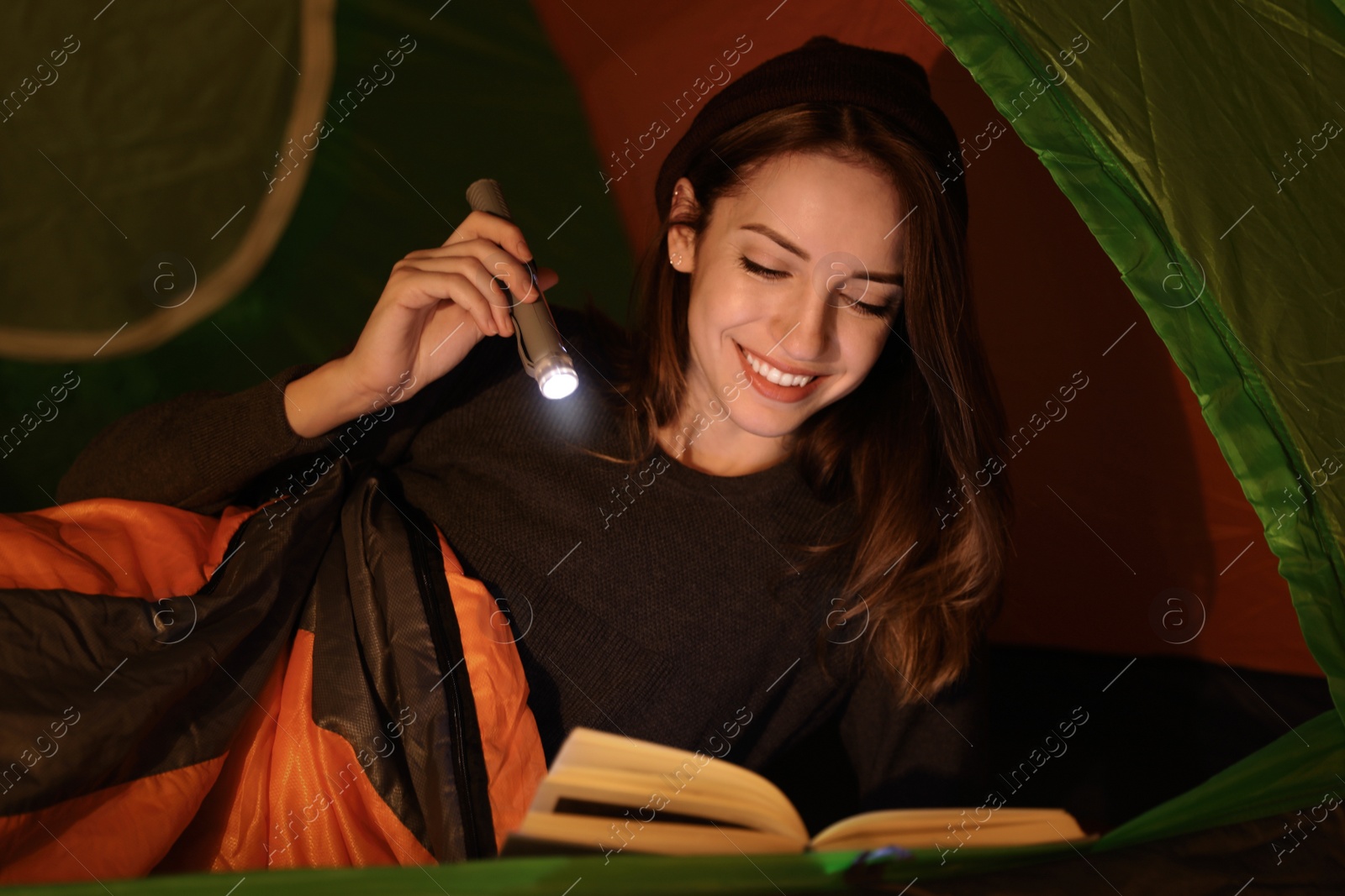 Photo of Young woman with flashlight reading book in tent