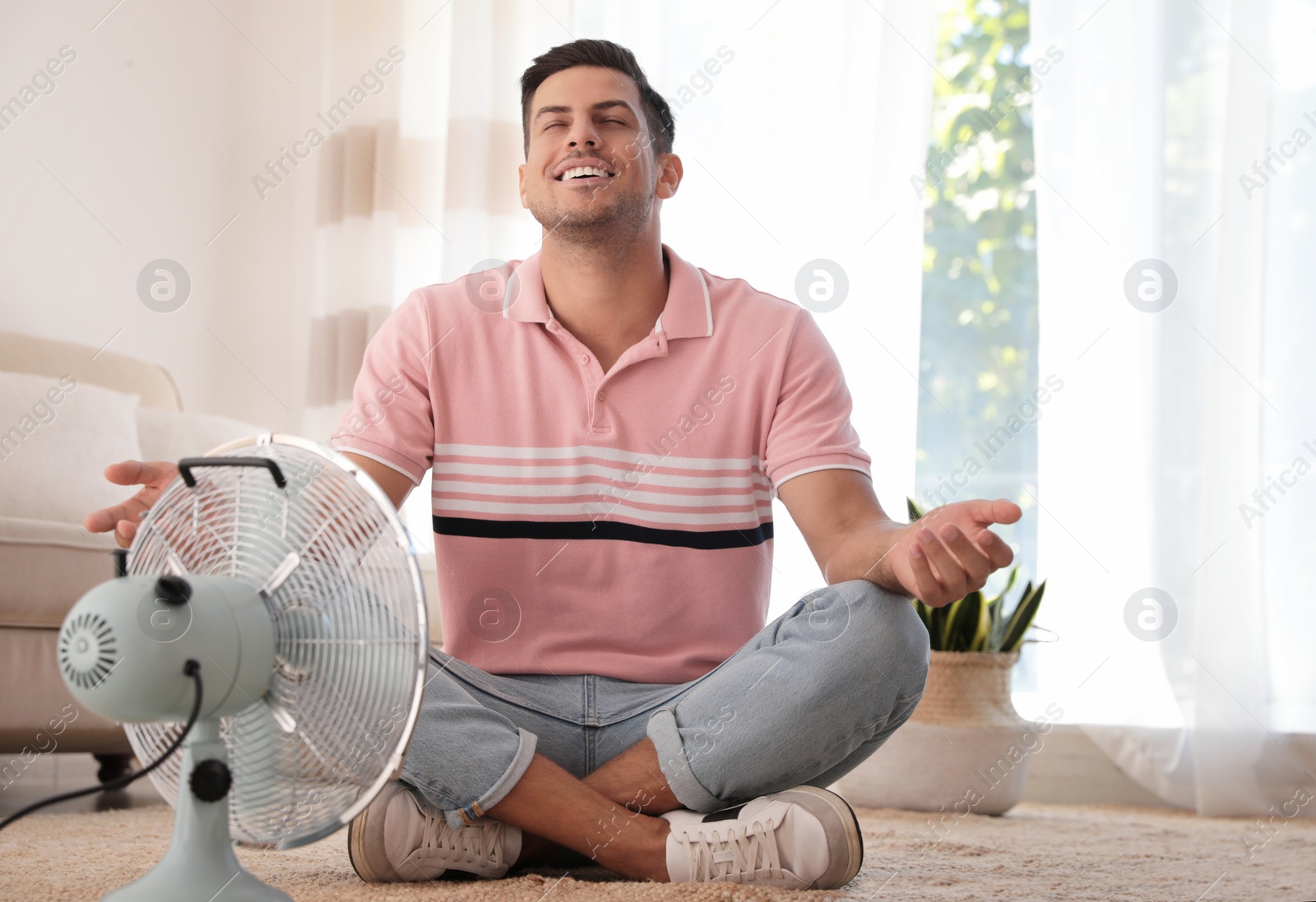 Photo of Man enjoying air flow from fan on floor in living room. Summer heat