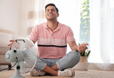 Photo of Man enjoying air flow from fan on floor in living room. Summer heat