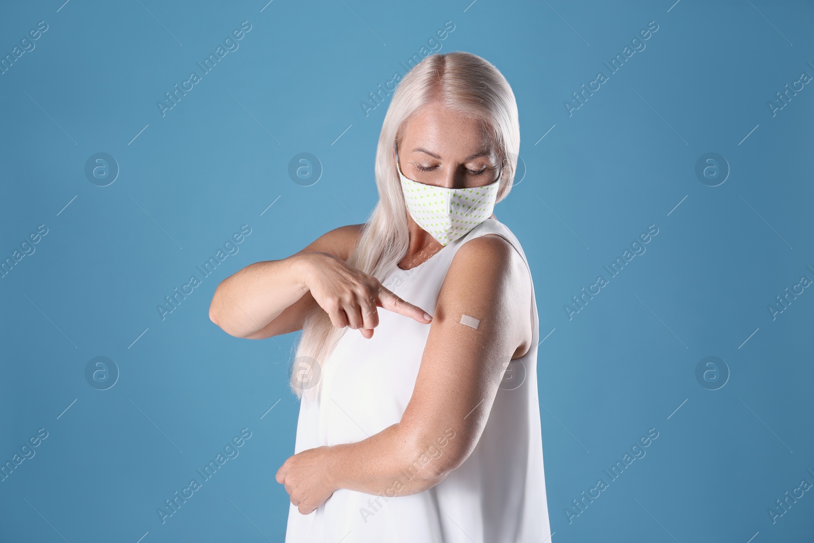 Photo of Mature woman in protective mask pointing at arm with bandage after vaccination on light blue background