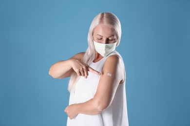 Photo of Mature woman in protective mask pointing at arm with bandage after vaccination on light blue background