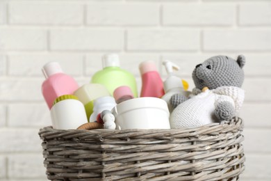 Wicker basket full of different baby cosmetic products and toys against white brick wall, closeup