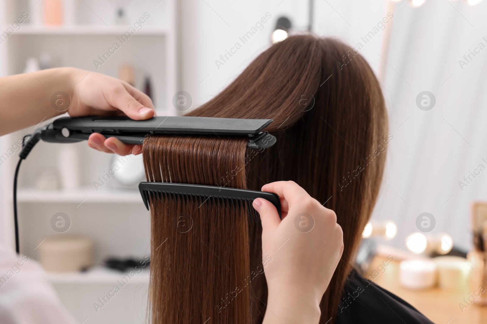 Photo of Hairdresser straightening woman's hair with flat iron in salon, closeup