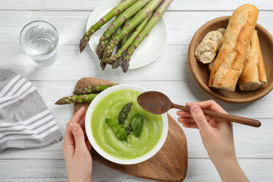 Woman eating delicious asparagus soup at white wooden table, top view