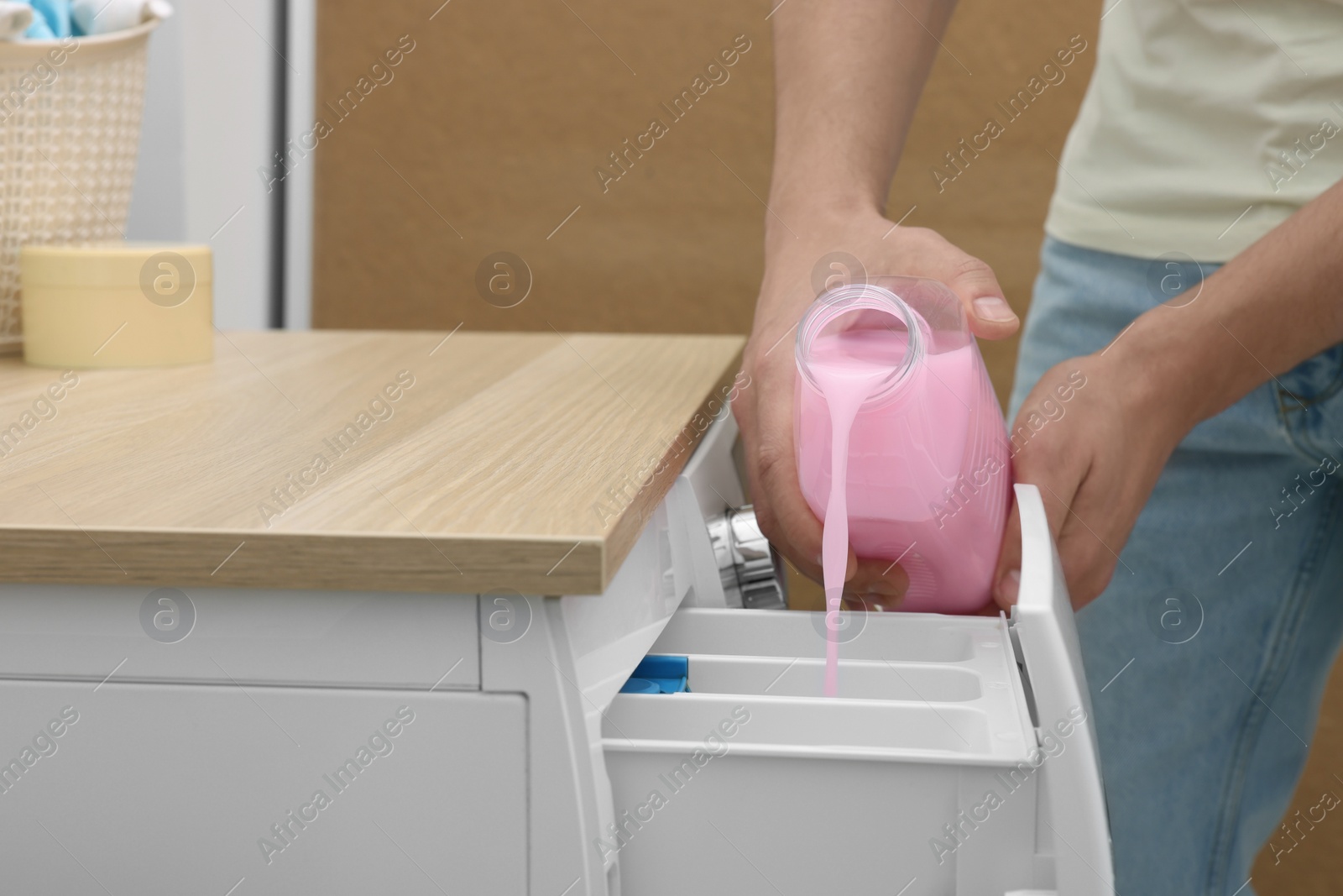 Photo of Man pouring fabric softener from bottle into washing machine indoors, closeup