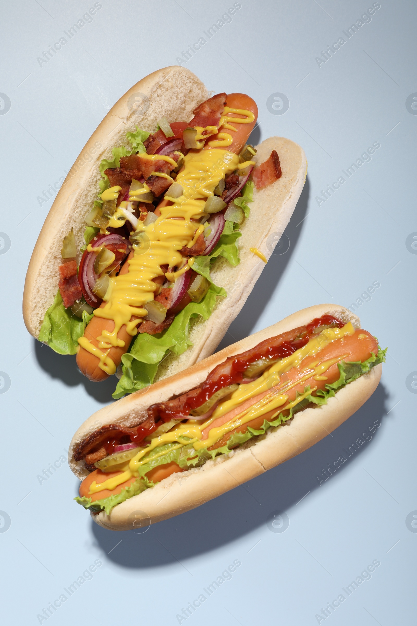 Photo of Delicious hot dogs with bacon, lettuce, onion and pickle on light blue background, flat lay