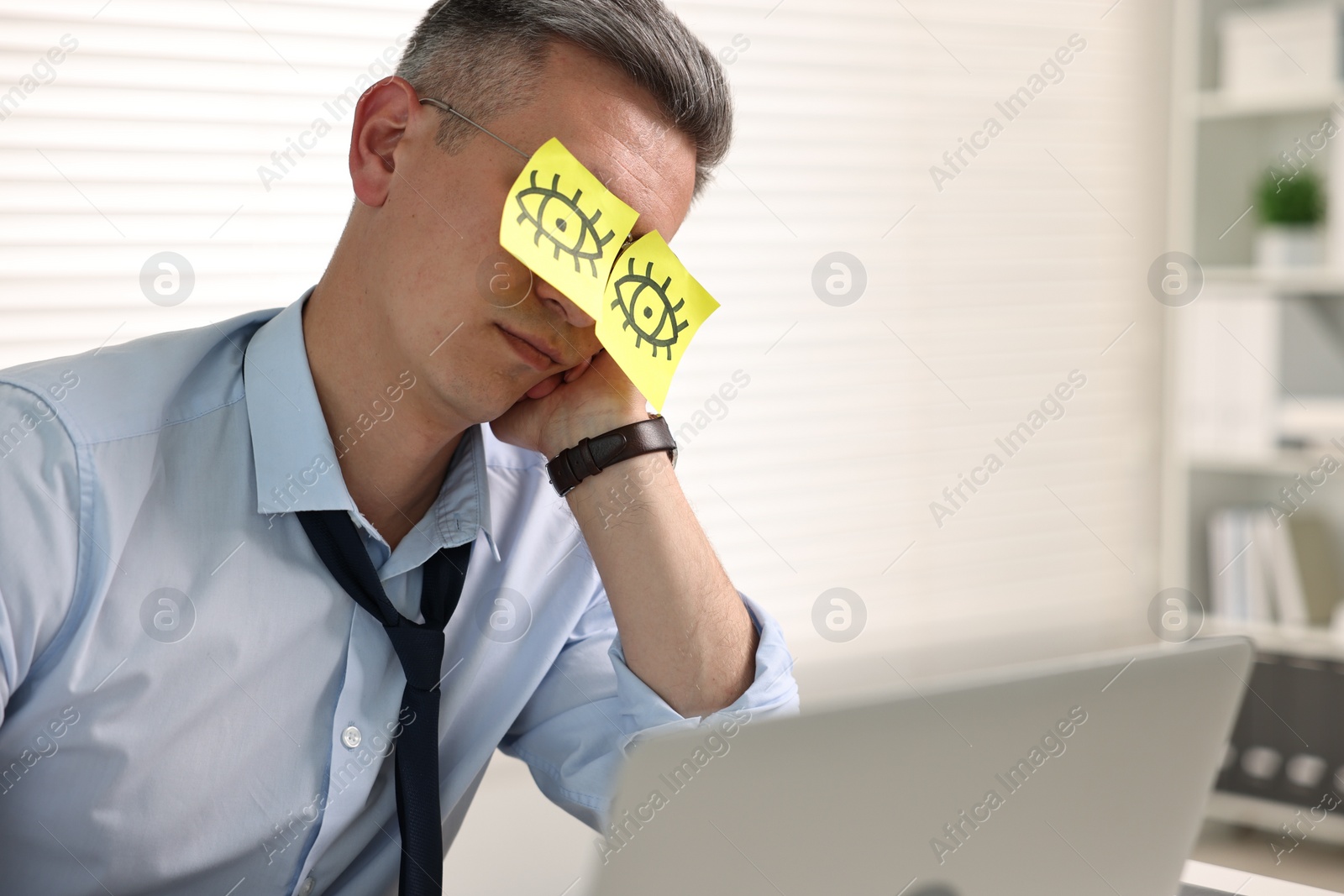 Photo of Man with fake eyes painted on sticky notes snoozing at workplace in office