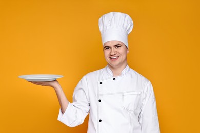 Photo of Portrait of happy confectioner in uniform holding empty plate on orange background