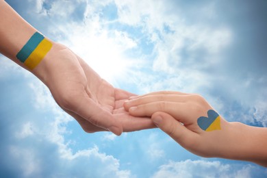 Image of Help for Ukraine. Woman and child holding hands with drawings of Ukrainian flag and beautiful blue sky on background, closeup