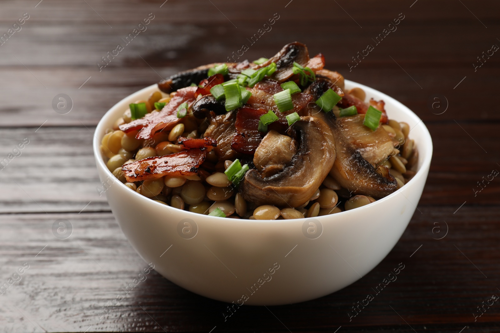 Photo of Delicious lentils with mushrooms, bacon and green onion in bowl on wooden table, closeup