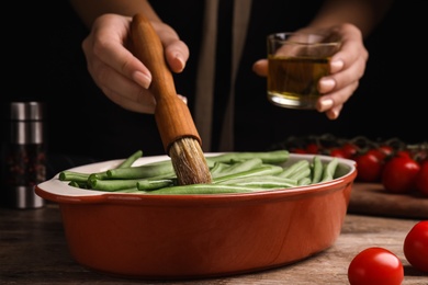 Woman brushing raw green beans with oil at wooden table, closeup