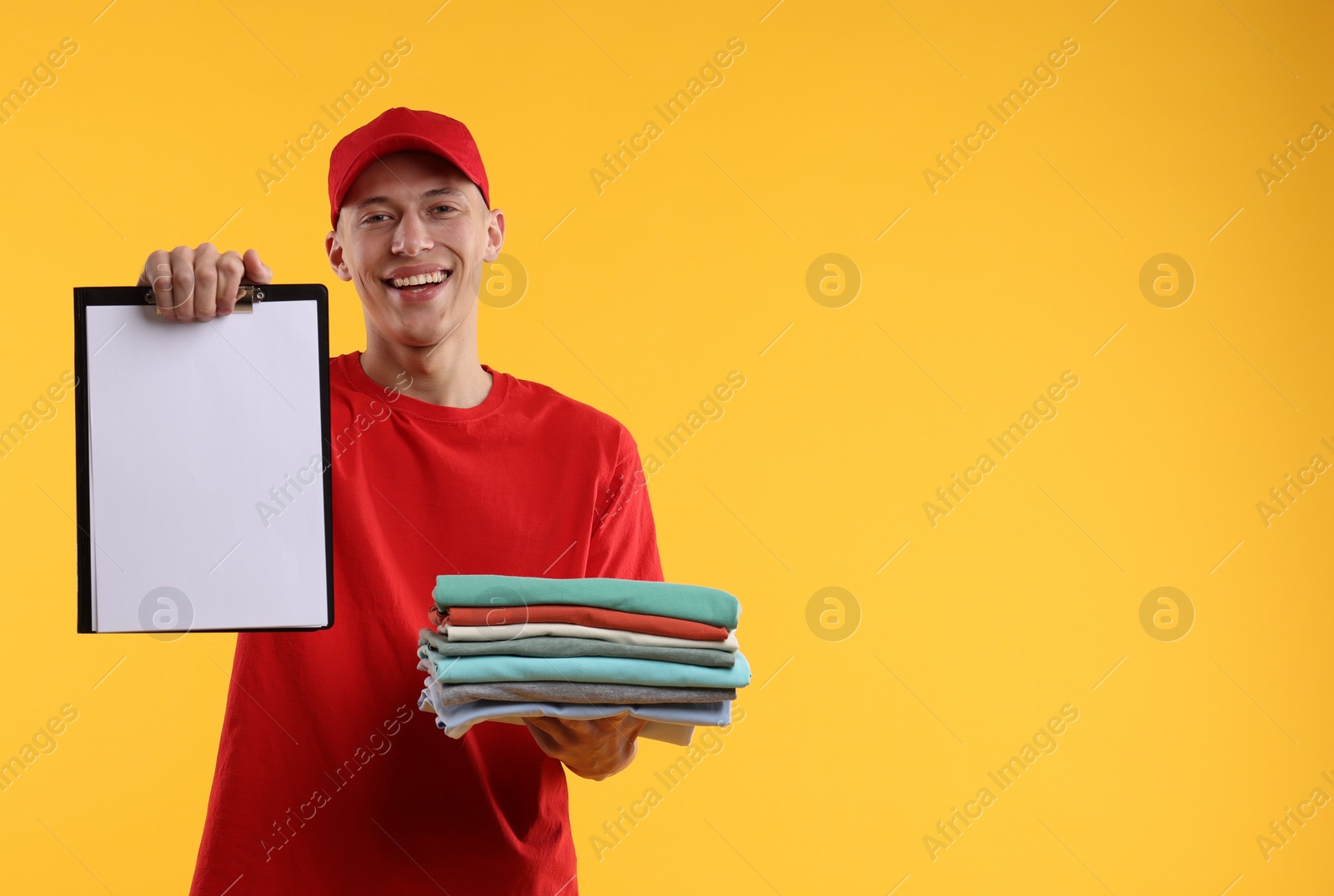 Photo of Dry-cleaning delivery. Happy courier holding folded clothes and clipboard on orange background, space for text