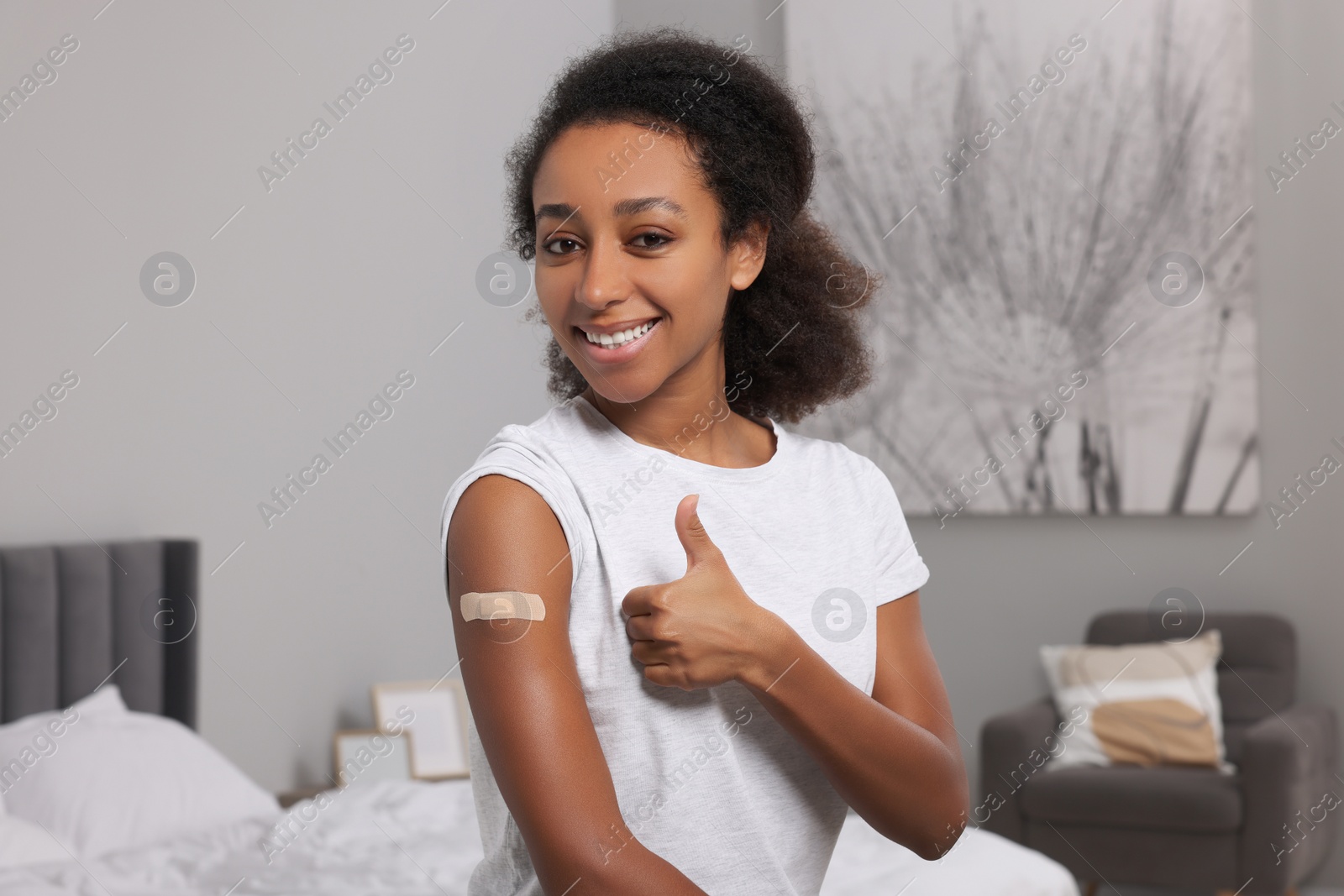 Photo of Happy young woman with adhesive bandage on her arm after vaccination showing thumb up indoors
