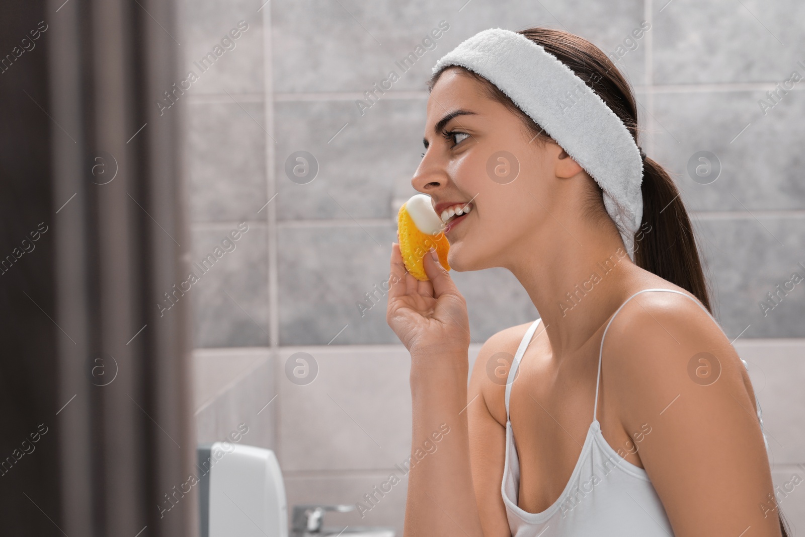 Photo of Young woman with headband washing her face using sponge in bathroom