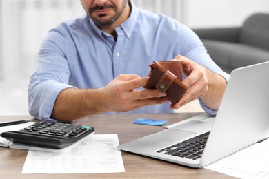 Photo of Man with empty wallet, laptop and calculator planning budget at table indoors, closeup. Debt problem