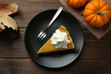 Piece of delicious pie with whipped cream, pumpkins and fork on wooden table, flat lay